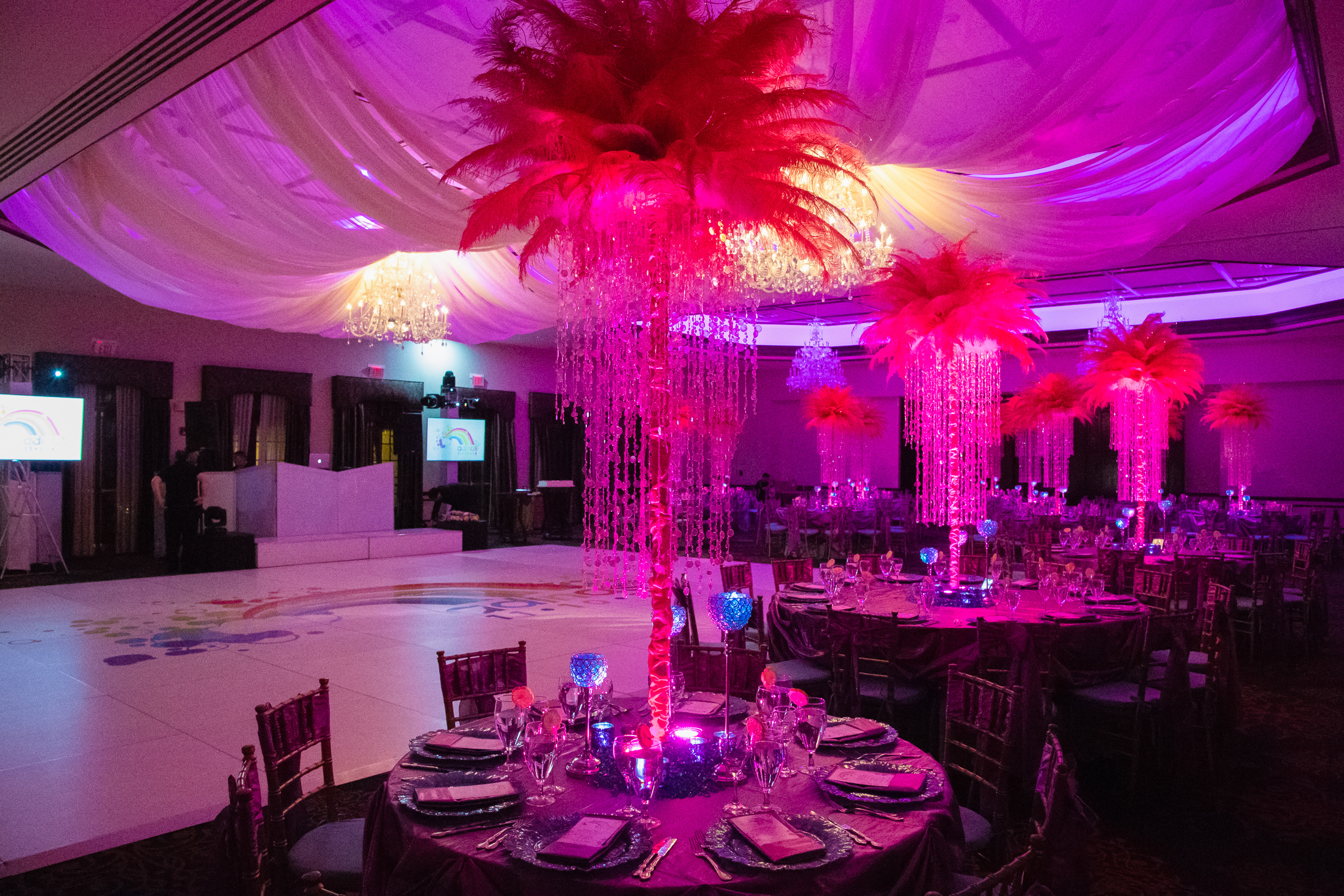 Mitzvah ballroom with pink lighting, draped ceiling, and feather centerpieces