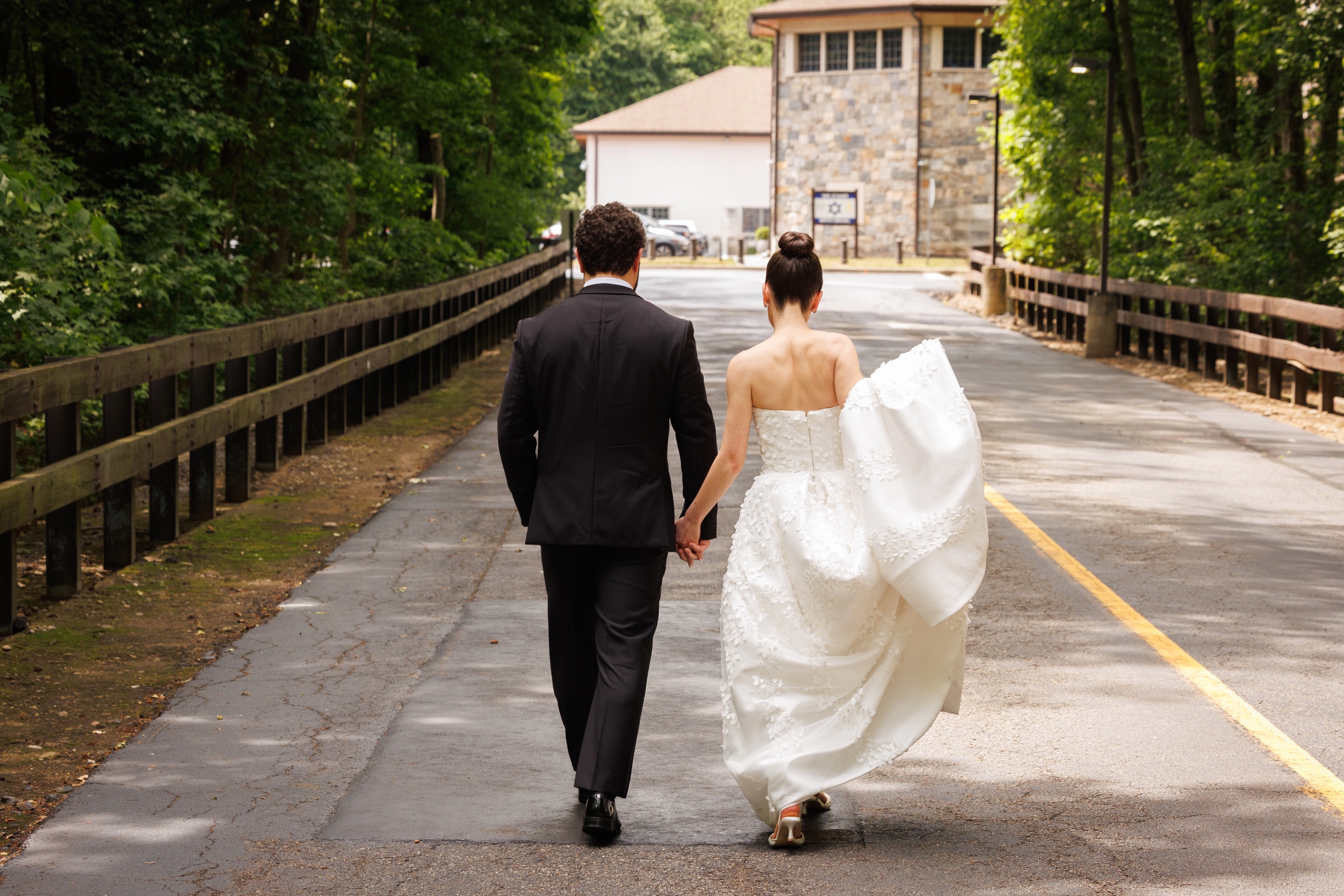 Newlyweds walking hand in hand toward the venue through the wooded grounds