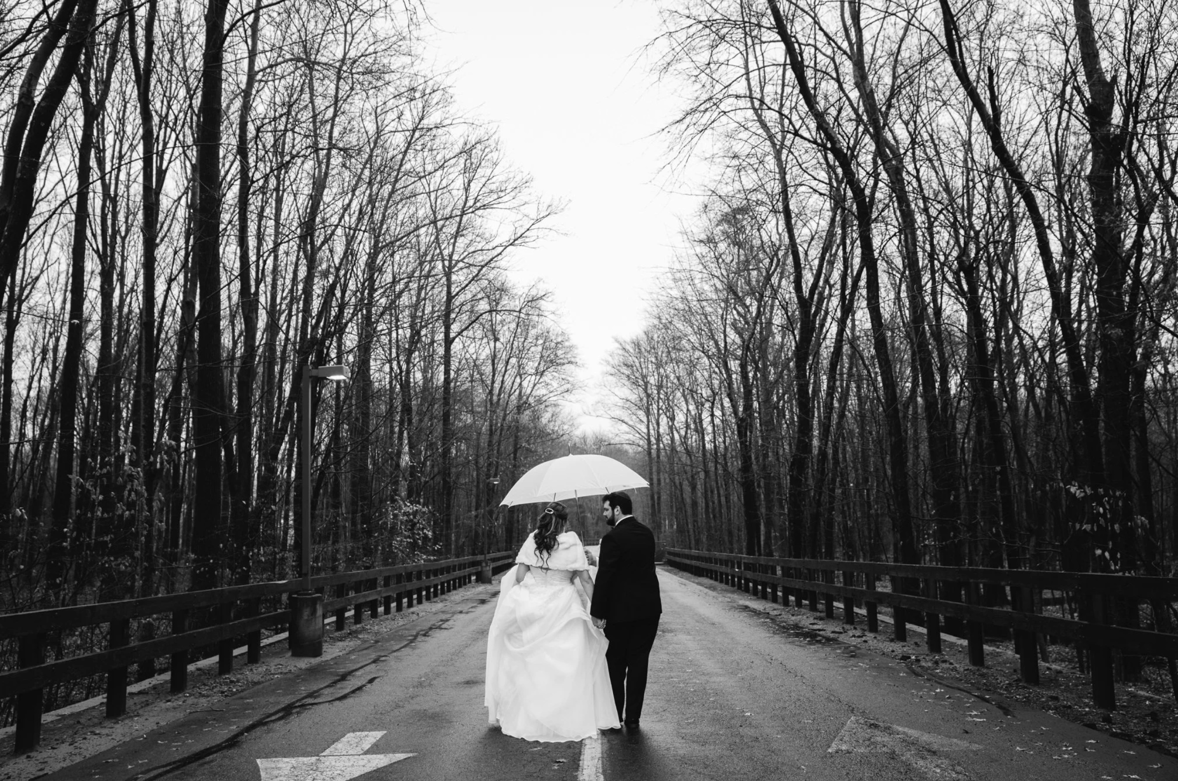 Black-and-white wedding portrait under an umbrella on a wooded road